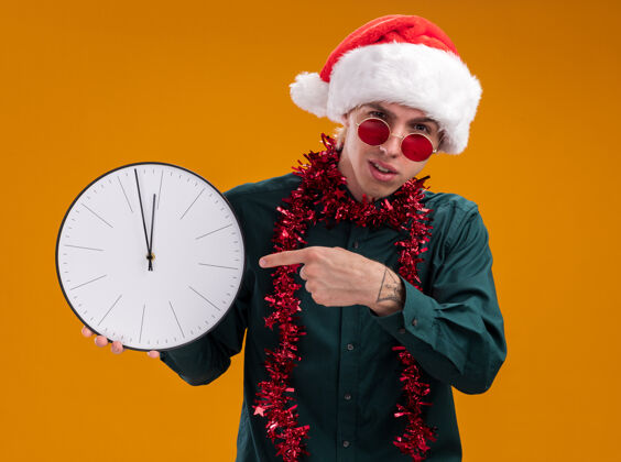 帽子皱眉的年轻金发男子戴着圣诞帽 戴着眼镜 脖子上戴着金属丝花环 拿着并指着时钟 看着孤立在橙色背景上的摄像机围着皱眉圣诞老人