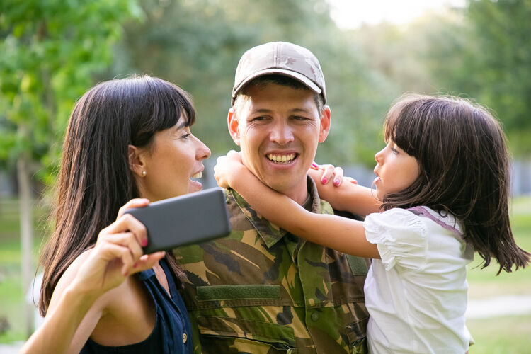 公园快乐的军人带着妻子和小女儿在城市公园里用手机自拍正面视图家庭团聚或回家的概念拍摄视图正面