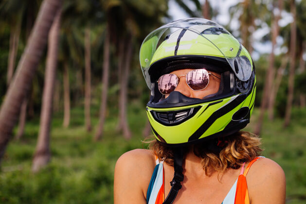 旅游在棕榈树下热带田野的丛林中 穿着黄绿色摩托车头盔和彩色浅夏装的美丽骑手妇女的肖像度假骑手异国情调