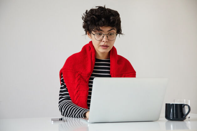 时尚困惑的年轻可爱的卷发黑发女人坐在桌边 带着现代笔记本电脑 一边扬起眉毛 一边看着屏幕 困惑的脸 隔离在白色背景下室内杯子毛衣