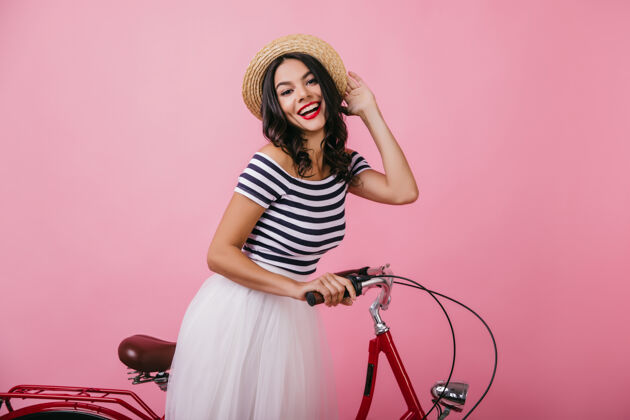 优雅优雅的拉丁女人穿着条纹t恤摆出情绪化的姿势漂亮的黑发女孩戴着帽子和自行车站在一起卷发女性模型
