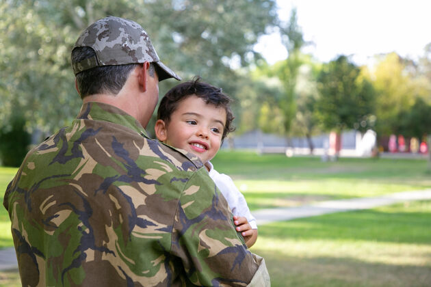 拥抱高加索人抱着孩子 穿着军装快乐的小男孩坐在父亲的手上 拥抱着父亲 开心地微笑家庭团聚 做父亲和回家的概念孩子军队父亲