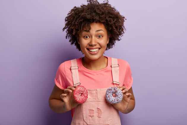 饮食乐观快乐的黑皮肤女人 非洲发型 拿着两个甜甜的油炸圈饼 享受甜食的乐趣乐观甜美非洲