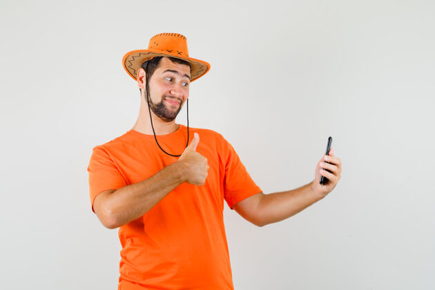 休闲穿着橙色t恤 戴着帽子 看起来很开心的年轻人一边自拍一边竖起大拇指正面图肖像衬衫年轻