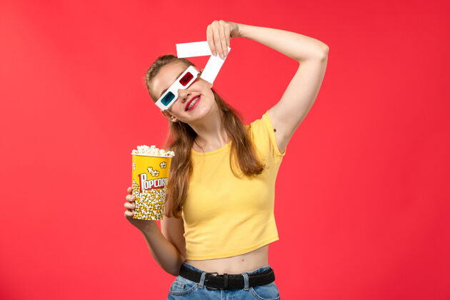 爆米花正面图年轻女子在电影院拿着爆米花包 戴着d型太阳镜 拿着红墙电影院的票 电影女孩身体电影院年轻的女性