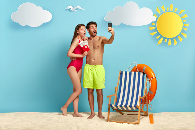 度假胜地令人惊讶的男女情侣在度假胜地享受暑假的水平镜头在手机摄像机前展示护照和登机票在蓝色背景的海滩上自拍机票防晒霜智能手机