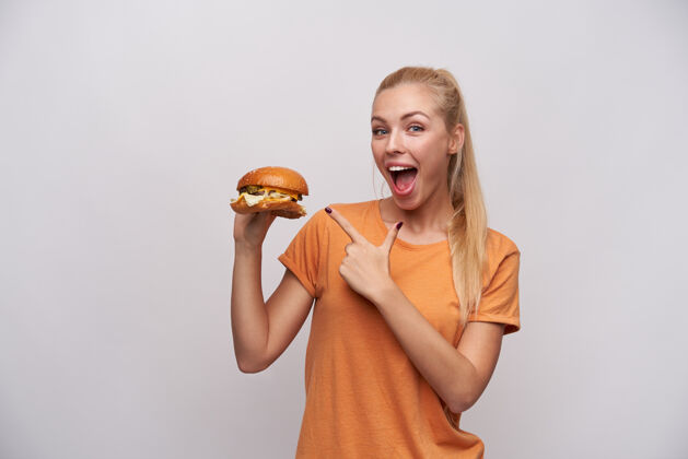 20多岁一位穿着休闲服的年轻金发女郎兴奋地抬起手 用食指指着新鲜的大汉堡 在白色背景下摆出一副开心的姿势 面带微笑地看着镜头金发漂亮表情