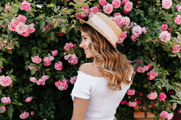 户外可爱的金发女孩戴着夏天的帽子微笑着看着花高兴的卷发女人放松与玫瑰拍照金发休闲夏天