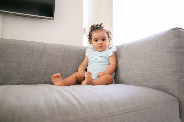 赛车严肃的深色卷发女婴穿着淡蓝色的布 坐在家里的灰色沙发上 一个孩子在家和童年的概念蝴蝶结低角度卷发