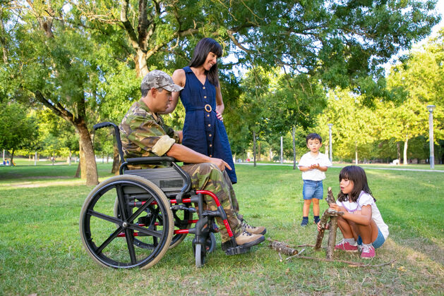 受伤快乐的妈妈和残疾的军人爸爸坐在轮椅上看着孩子们为户外篝火排柴火残疾退伍军人或家庭户外概念柴火孩子父母