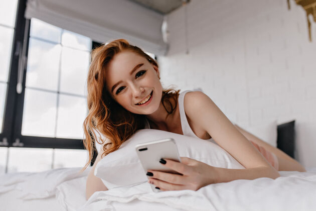 红发迷人的年轻女子 黑色美甲躺在床上微笑可爱的姜女摆在卧室里与智能手机成人积极年轻