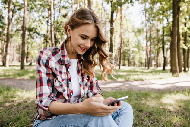 成人好奇的卷发女孩坐在草地上发短信美丽的时尚女士在森林里的户外照片森林智能手机欢快
