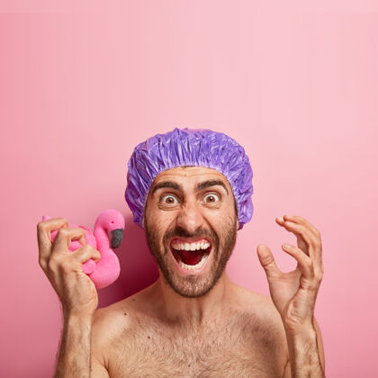 男情绪激动的男人积极地打手势 恼火地拿着柔软的粉红色火烈鸟海绵 戴着浴帽凝视身体护理男性