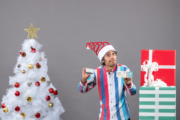年轻人前视图：年轻人拿着圣诞树和礼物周围的卡片和旅行票 目光敏锐人树周围