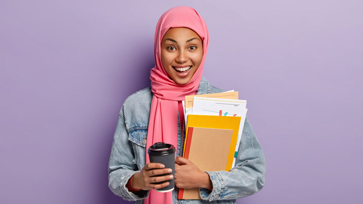 笔记本快乐的少数民族女学生有咖啡休息时间 拿着外卖杯饮料 拿着笔记本和纸 头上戴着粉色围巾 伊斯兰宗教观点 在室内摆姿势人 文化 传统传统大学学习