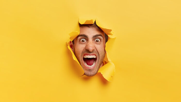 水平愤怒的男人脸穿过黄色的纸洞愤怒的男人把头伸过撕破的背景尖叫反应昏迷