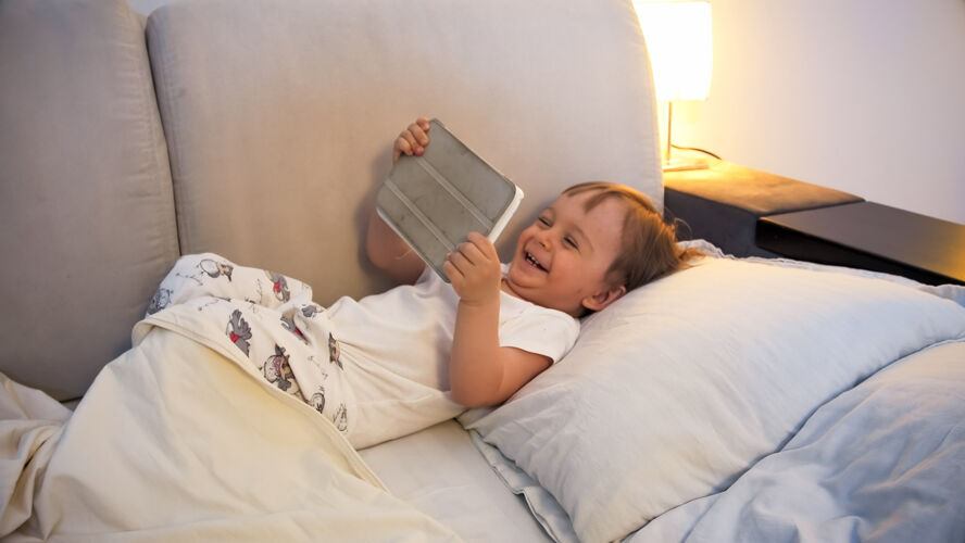 小快乐的欢笑的学步男孩晚上躺在床上 在数字平板电脑上看动画片乐趣父母宝宝