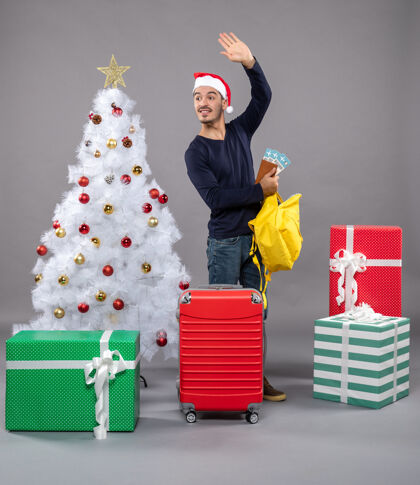盒子一个戴着圣诞帽 带着红色手提箱的年轻人在不同的礼物周围检查他的黄色背包手提箱帽子圣诞树