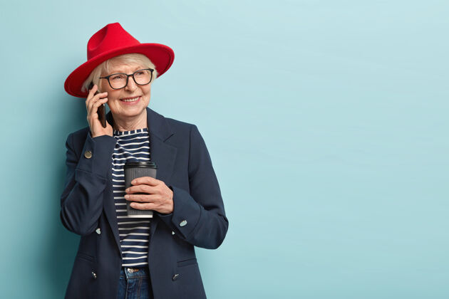 祖母人 年龄 休闲的概念快乐的老太太享受业余时间 有电话交谈 喝咖啡去 戴着红色的头饰和正式的外套 看旁边 模型在蓝色的墙壁 自由的空间饮料眼镜眼镜