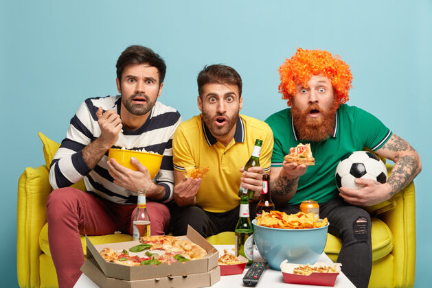 足球兴奋的三个男朋友看体育比赛 在危险时刻屏住了呼吸 咬着薯片爆米花和披萨坐在客厅的黄色沙发上球迷们拿着啤酒足球支持者足球得分球迷