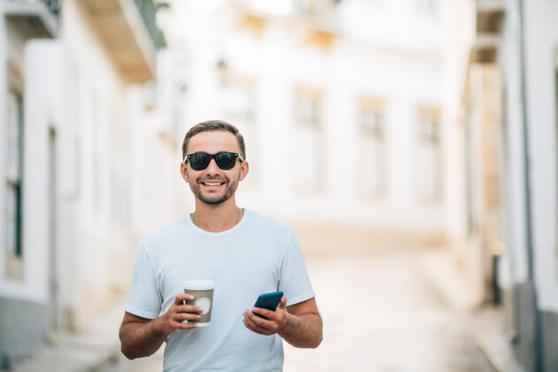 商务男士自信的年轻人戴着眼镜拿着咖啡杯 在户外散步时使用智能手机包专业风格