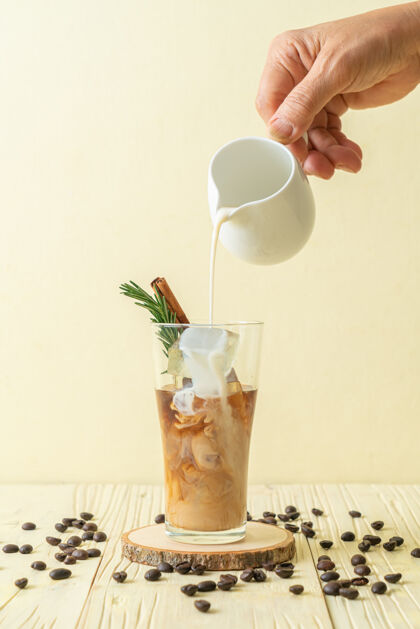 早晨把牛奶倒在黑咖啡杯里 把冰块 肉桂和迷迭香放在木桌上冷冻冰咖啡深色