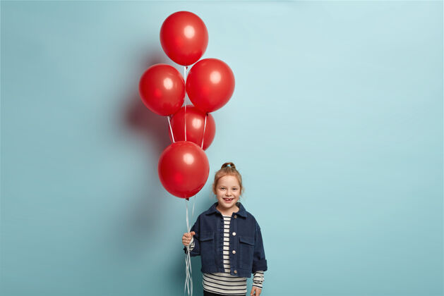 庆祝可爱的小男孩穿着时尚的牛仔夹克站在红色气球 在朋友生日到来 有快乐的表情 站在蓝色的墙壁童年和庆祝的概念气球小高兴