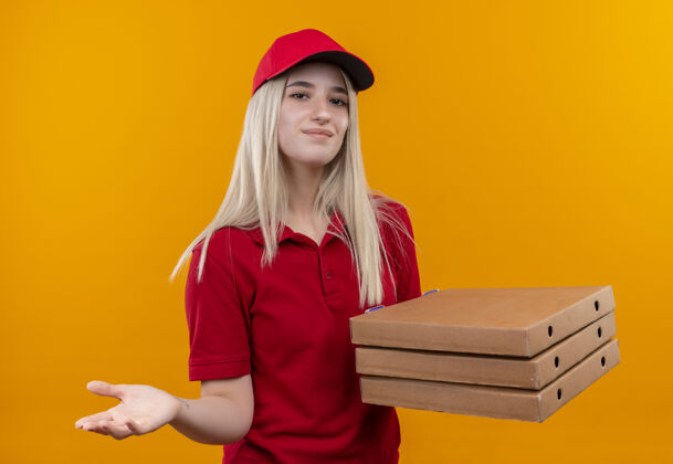 帽子在孤立的橙色背景下 一个穿着红色t恤 戴着帽子的年轻女孩拿着比萨饼盒向镜头伸出手来送货高兴年轻