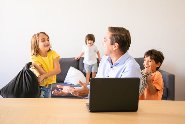 女儿高加索人的爸爸和顽皮的孩子们聊天 坐在桌子旁快乐的中年爸爸在家里用笔记本电脑和孩子们玩枕头童年和数字技术的概念可爱童年室内