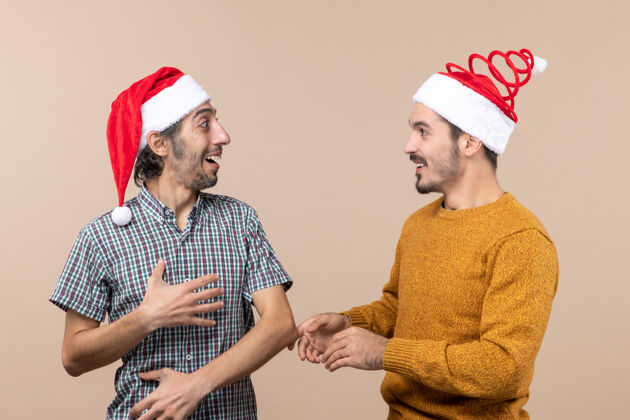 肖像正面图两个带着圣诞帽的微笑的家伙 一个在米色孤立的背景下看着对方米色帽子帽子