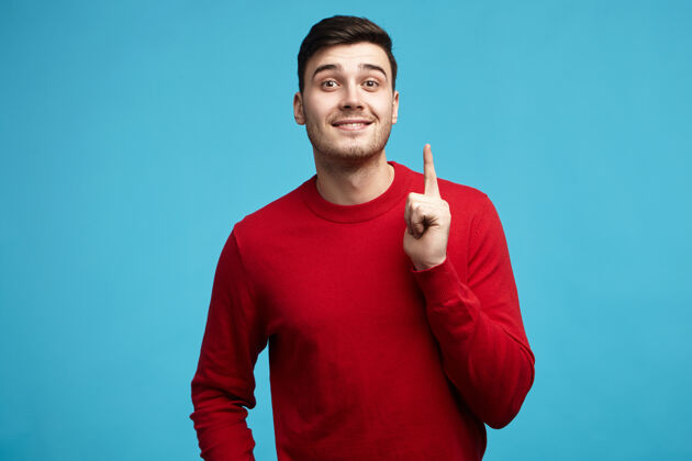 男人情绪化的未刮胡子的欧洲年轻人穿着红色毛衣举起食指的照片吸引你的注意手成人胡须