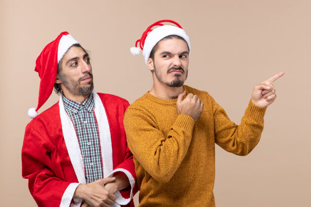 显示前视图两个戴着圣诞帽的朋友一个看着另一个 另一个在米色孤立背景上显示方向两个前面方向