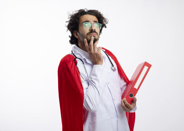 手体贴的年轻白种人超级英雄戴着眼镜 身穿医生制服 披着红色斗篷 脖子上戴着听诊器 手放在下巴上 手里拿着文件夹和复印件眼镜男人医生
