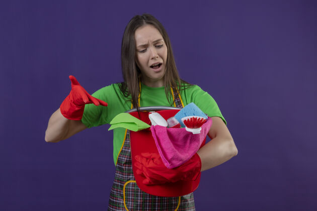 紫悲伤的清洁年轻女孩穿着制服 戴着红色手套 指着她手上的清洁工具 背景是孤立的紫色制服女孩年轻