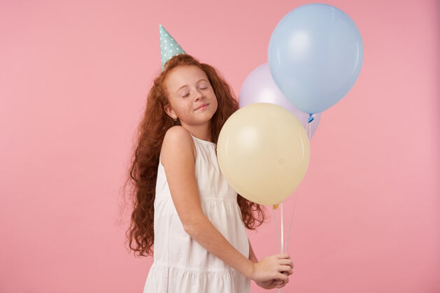 快乐美丽微笑的小女孩 一头浓密的卷发 穿着优雅的连衣裙 戴着生日帽 在粉色背景下 用气球摆出姿势 在庆祝节日的同时表达真正的积极情绪惊喜女孩积极