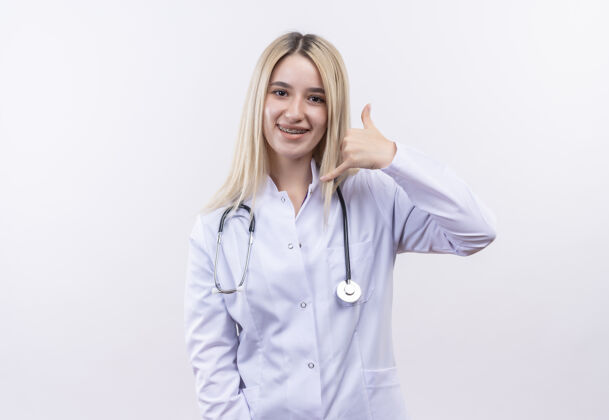 手势微笑的医生 年轻的金发女孩 戴着听诊器和医用长袍 戴着牙套 在孤立的白色背景上显示呼叫手势金发微笑女孩