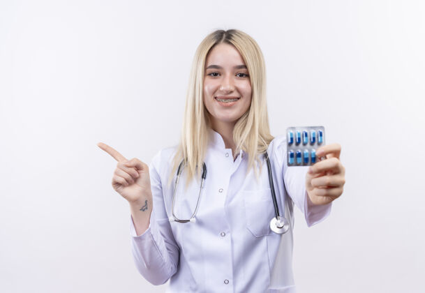 医疗微笑的医生年轻的金发女孩戴着听诊器和医用长袍 戴着牙套 拿着药片 在孤立的白色背景上指向一边点听诊器侧边
