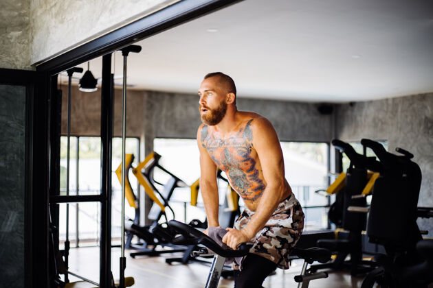 年轻人纹身 肌肉发达 胡须浓密的男人骑着自行车在体育馆里锻炼有氧运动 靠近大窗户 外面可以看到树训练努力运动装