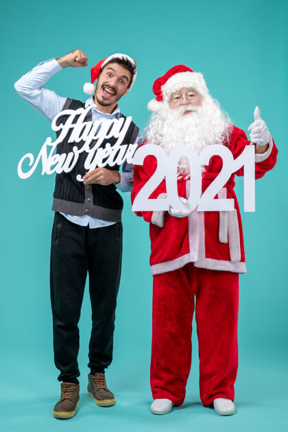 十二月圣诞老人的正面图 男性手持新年快乐和2021年板在蓝色墙上圣诞老人乐趣服装