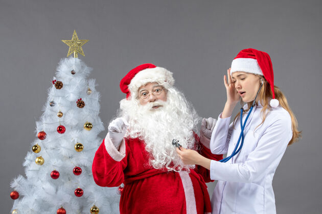快乐圣诞老人和女医生的前视图 女医生正在灰墙上检查他的健康状况庆祝健康服装