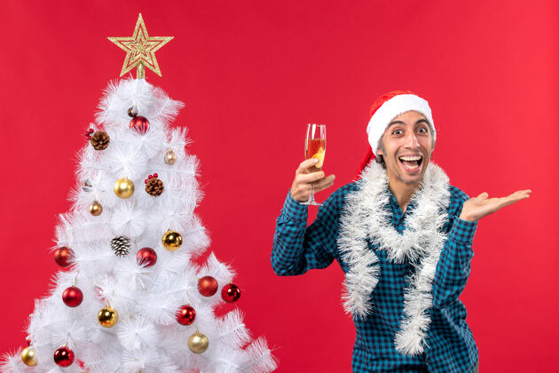 男人圣诞心情带着微笑快乐的年轻人 戴着圣诞老人帽 穿着蓝色条纹衬衫 在圣诞树旁拿着一杯葡萄酒装饰庆祝玻璃