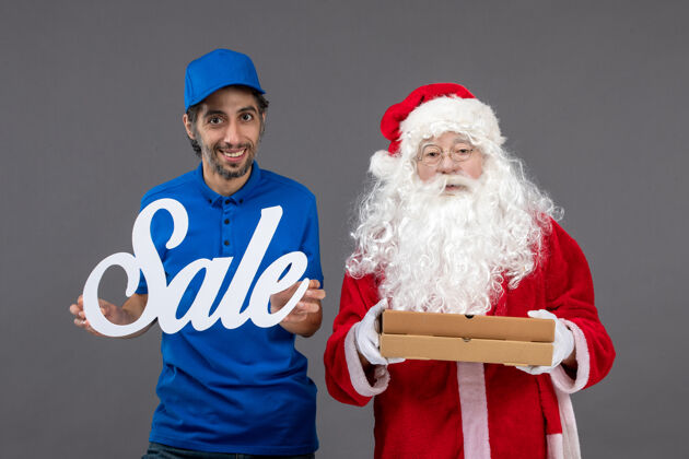 十二月圣诞老人的正面图 男信使手持销售横幅 灰色墙上有食品盒服装信使人