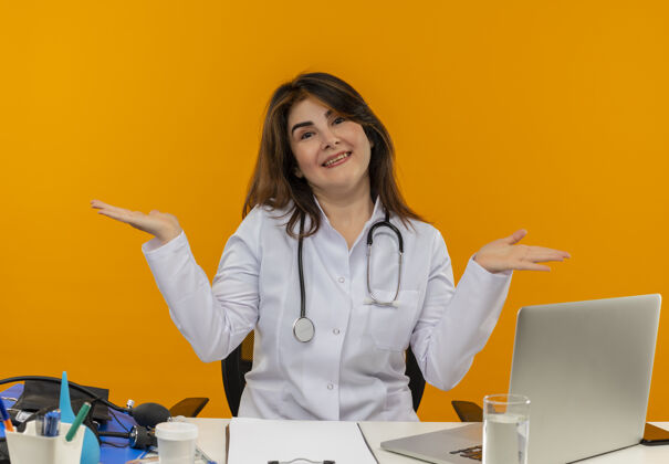 医生微笑的中年女医生穿着带听诊器的医用长袍 坐在办公桌旁 拿着医疗工具在笔记本电脑上工作 双手放在隔离的橙色背景上 还有复印空间中年穿着女性
