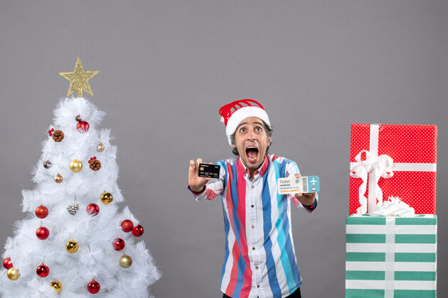 无辜前景：一个拿着卡片和旅行票的男人高高兴兴地看着圣诞树和礼物卡片帽子男性