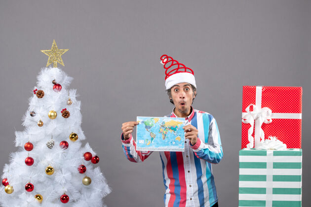 举行前视图惊讶的男子与螺旋弹簧圣诞帽和条纹衬衫举行地图附近的白色圣诞树和复制空间人和服惊喜的人