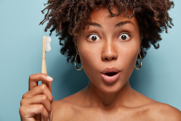 牙齿惊讶的黑皮肤女人早上执行日常卫生程序 震惊的是几乎没有时间准备 赶紧上班 拿着木制牙刷 半裸在浴室里摆姿势惊讶凝视牙齿