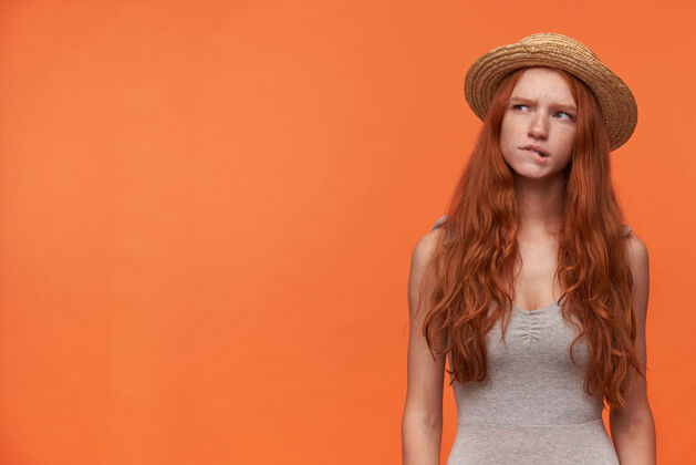 女士摄影棚照片中 穿着休闲服的年轻红发美女站在橙色背景下 身穿灰色衬衫 头戴水手帽 若有所思地看着旁边 咬着屁股衬衫长帽子