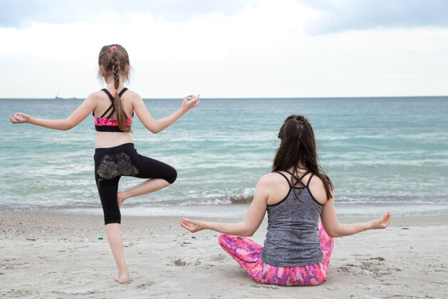 海滨妈妈带着小女儿穿着运动服在海边练习瑜伽 从后面看家庭价值观和健康的生活方式快乐的家庭家庭生活方式