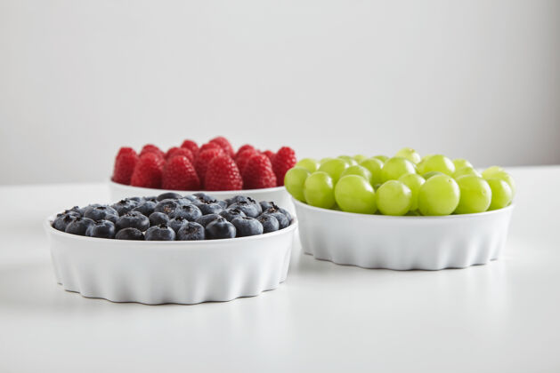 甜点一堆成熟的覆盆子 蓝莓和绿色无籽麝香葡萄准确地放在白色桌子上的陶瓷碗里健康抗氧化剂饮食