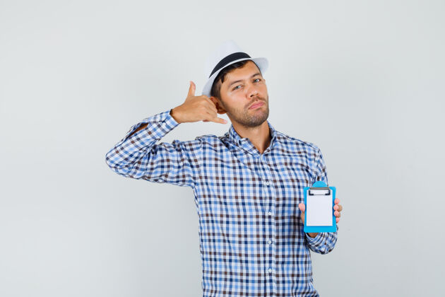 人年轻人拿着迷你剪贴板 在格子衬衫里展示电话手势成年人帽子人